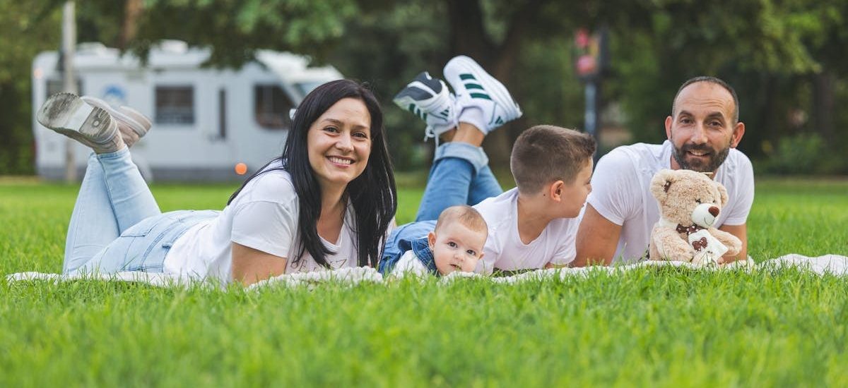 Happy family lying on grass with RV in the background, enjoying a sunny outdoor picnic.
