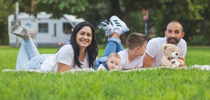Happy family lying on grass with RV in the background, enjoying a sunny outdoor picnic.